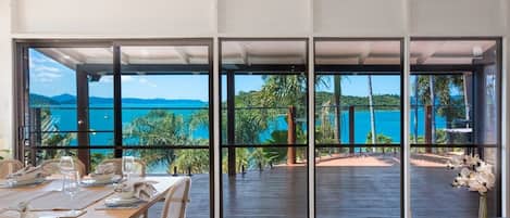 A dining area with a table set for two, large windows offering a view of the sea and greenery.