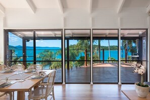A dining area with a table set for two, large windows offering a view of the sea and greenery. - Coral Sea Pearl (Shute Harbour)