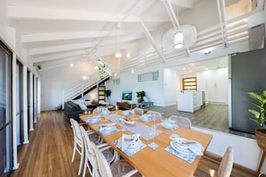 Un comedor moderno con una larga mesa de madera dispuesta para una comida, una escalera que lleva a un nivel superior, y vistas al área de la cocina.