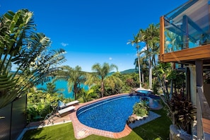 Une piscine à bord incurvé, entourée d’une végétation luxuriante et de palmiers, offrant une vue dégagée sur l’océan et les collines en arrière-plan.