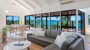 A spacious living area with a large window overlooking a tropical landscape, a leather sofa, and a dining area with natural light. - Coral Sea Pearl (Shute Harbour)