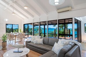 Amplia sala de estar con un gran ventanal con vistas a un paisaje tropical, un sofá de cuero y un comedor con luz natural.