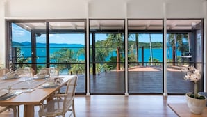 Un comedor con una mesa dispuesta para dos, ventanas amplias que ofrecen vistas a un paisaje tropical y el mar.