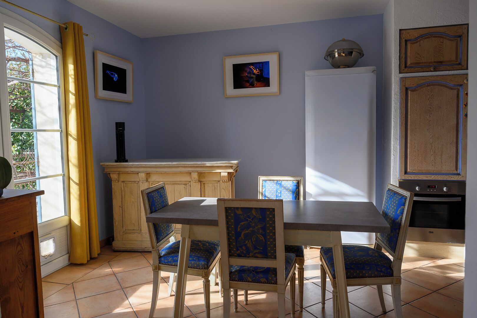 A dining area with a wooden table and blue upholstered chairs, a kitchen with wooden cabinets, and a window with yellow curtains.