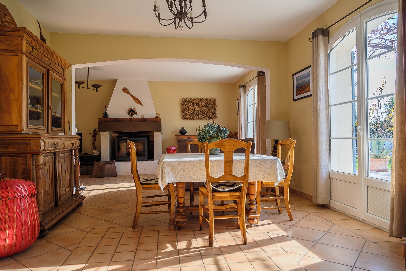 A dining area with a table set for four, wooden chairs, a fireplace, and a large window with curtains.
