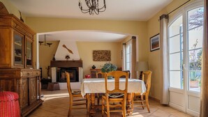 Un coin repas avec une table dressée pour quatre personnes, des chaises en bois, une cheminée et une grande fenêtre avec des rideaux.