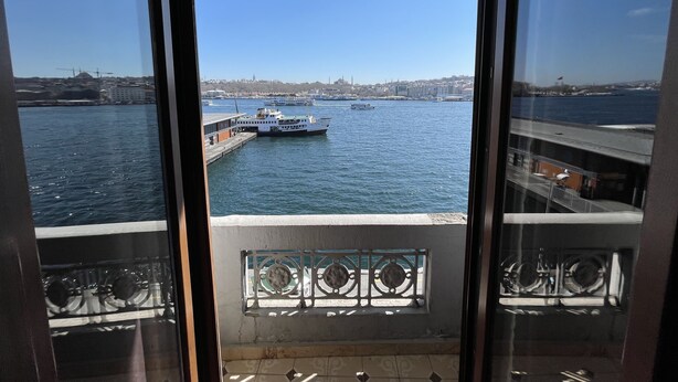A view from a window overlooking a body of water with boats, a cityscape in the background, and a balcony with a railing. - Weingart Seaside Hotel (Istanbul)