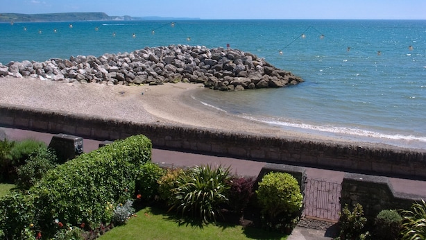 A coastal view with a rocky breakwater, a paved walkway, and lush greenery. - Weymouth Bay Apartment A (Weymouth)