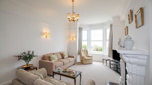 A spacious living room with a beige sofa set, a coffee table, a fireplace, and a large window with curtains. - Weymouth Bay Apartment A (Weymouth)