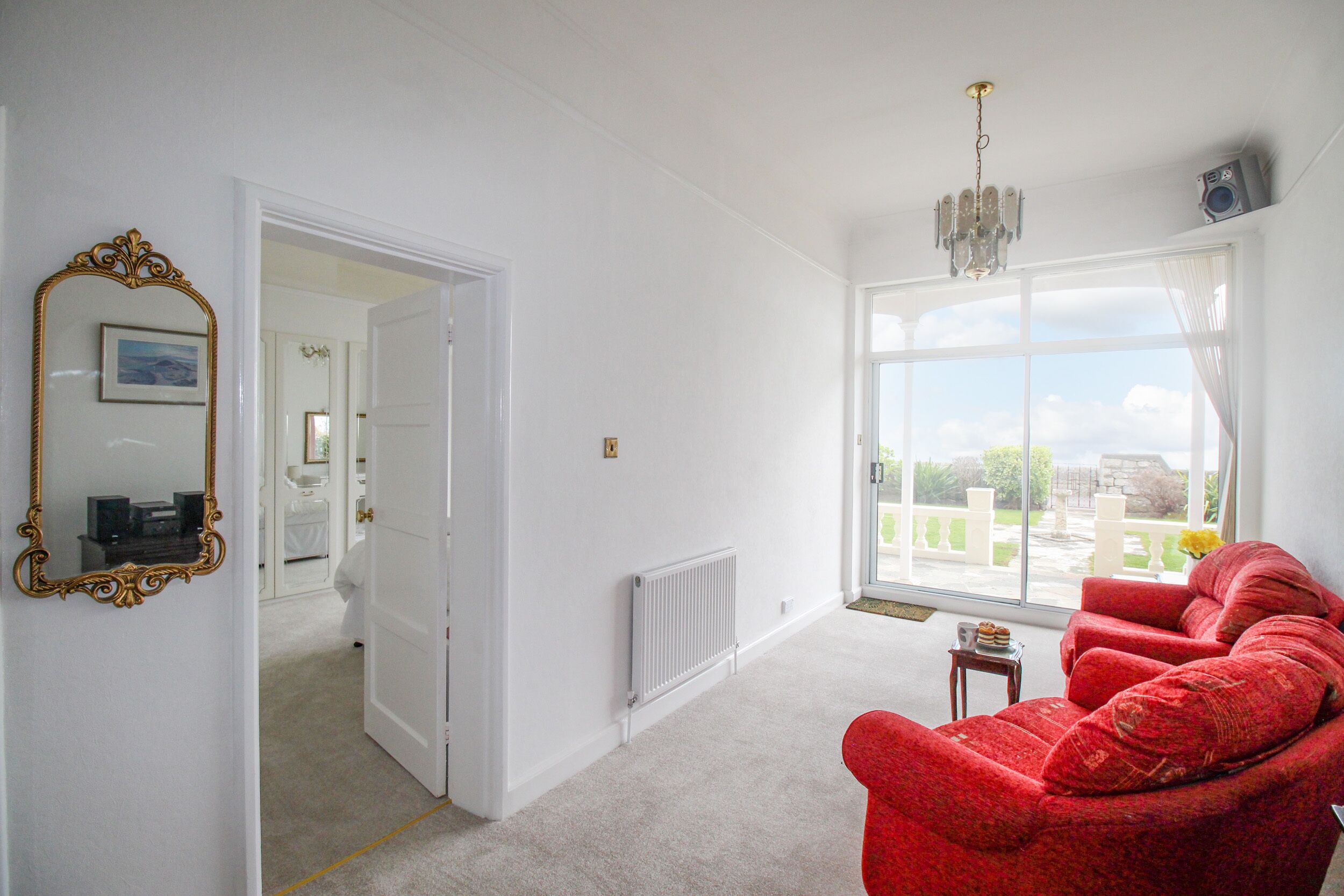 Una habitación amplia con un sillón rojo, un ventanal con vistas, un radiador y una puerta que da a otra habitación.