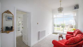 Una habitación amplia con un sillón rojo, un ventanal con vistas, un radiador y una puerta que da a otra habitación.