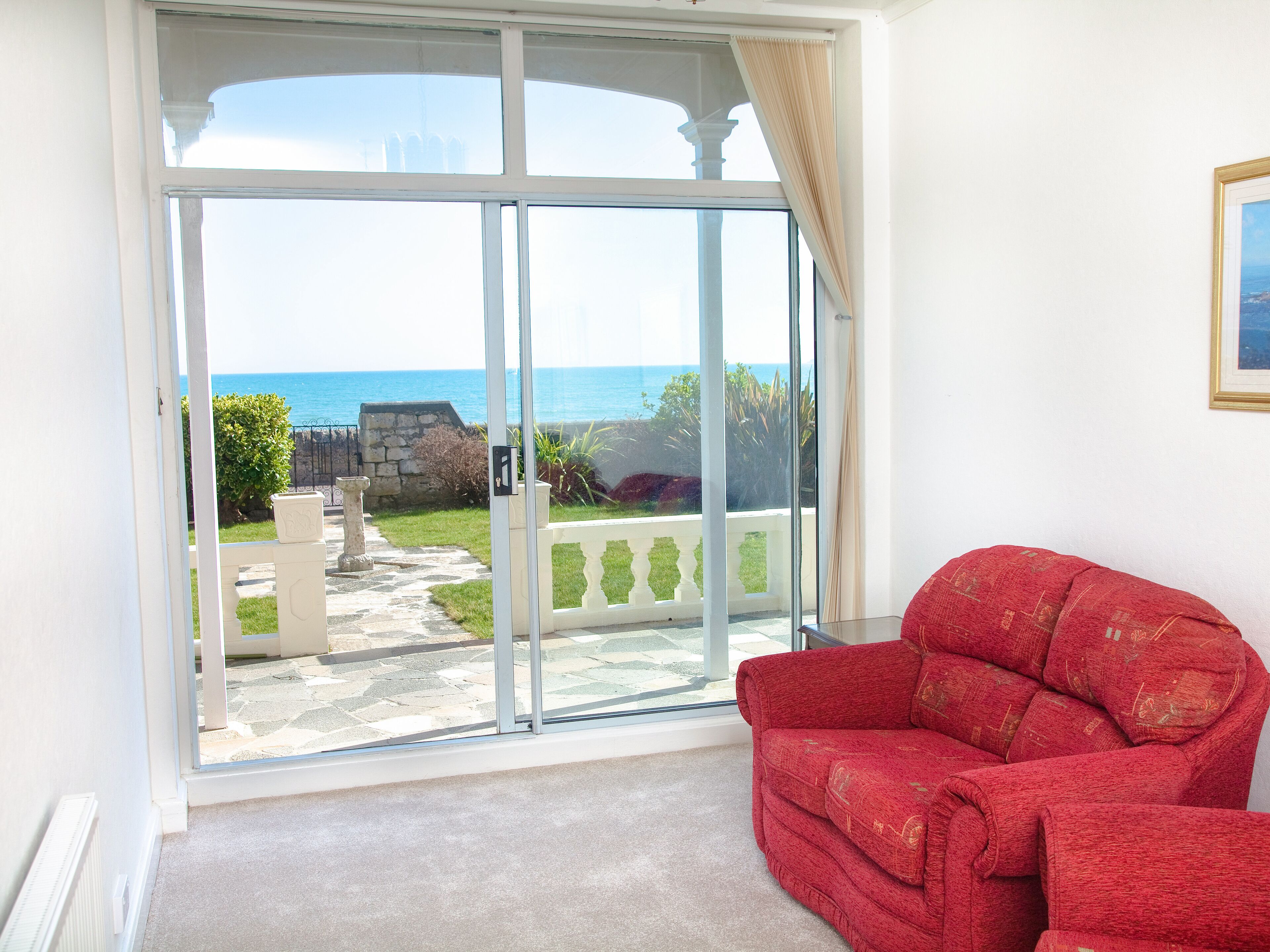 Um quarto com um sofá vermelho, uma grande porta de correr de vidro que dá para uma varanda com vista para o mar, e um quadro pendurado na parede.