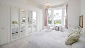 A bedroom with a large bed, a chandelier, a window with curtains, a chair, and a mirror. - Weymouth Bay Apartment A (Weymouth)
