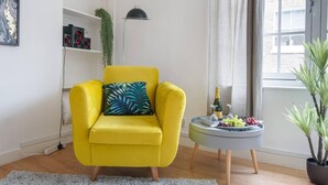 A yellow armchair with a leaf-patterned pillow, a small side table with a bottle and grapes, and a potted plant.