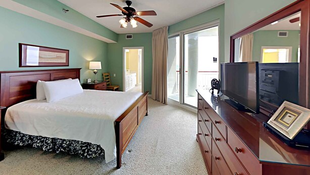 A hotel room with a bed, a dresser, a television, and a ceiling fan. - Spacious Gulf Front Condo - Stunning Views + On-Site Amenities! (Navarre Beach)