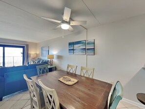 A dining area with a wooden table, four chairs, a ceiling fan, and a view of the ocean through a large window.
