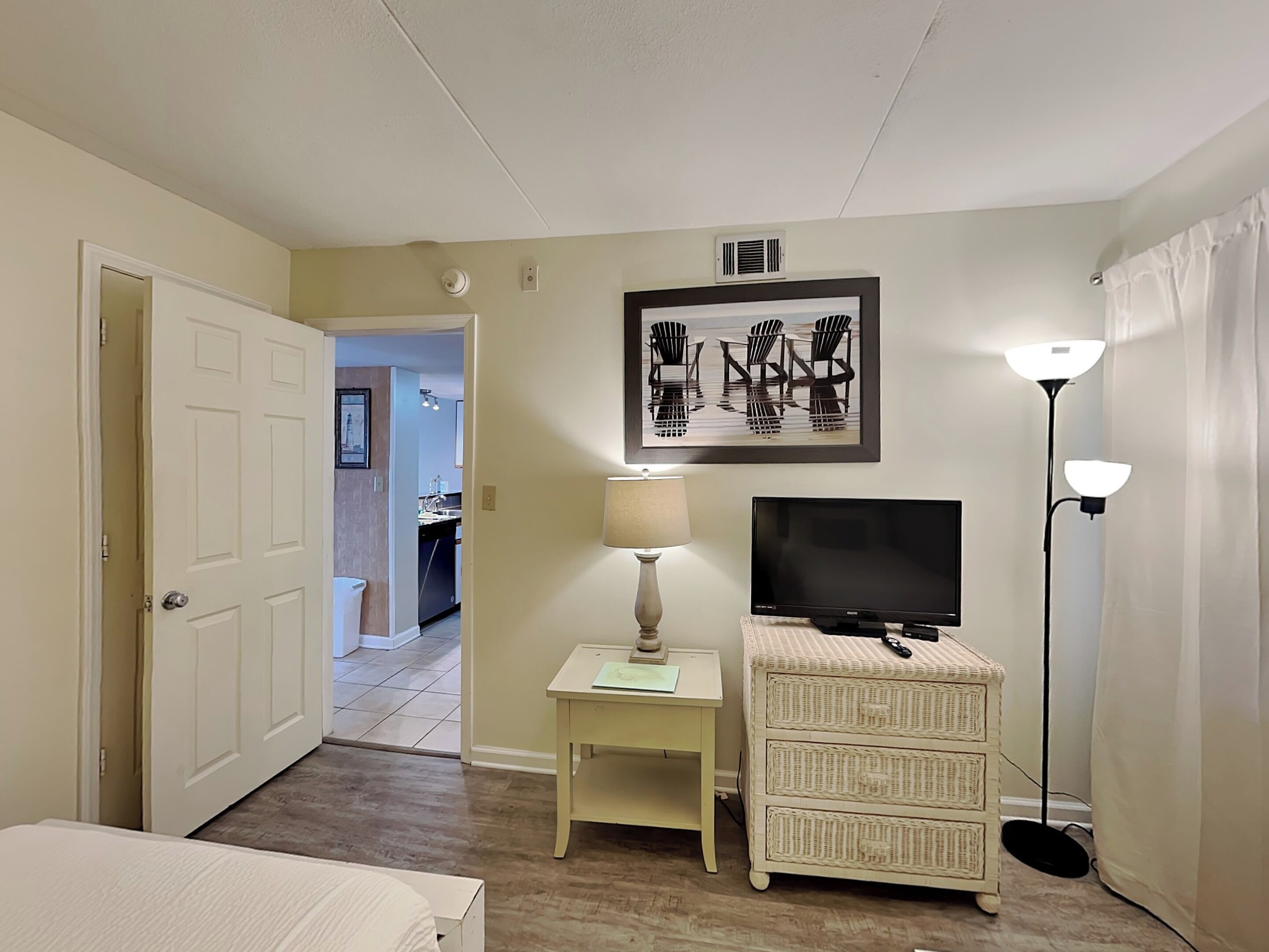 A hotel room with a bed, a TV, a lamp, a wicker chest of drawers, and a painting on the wall.