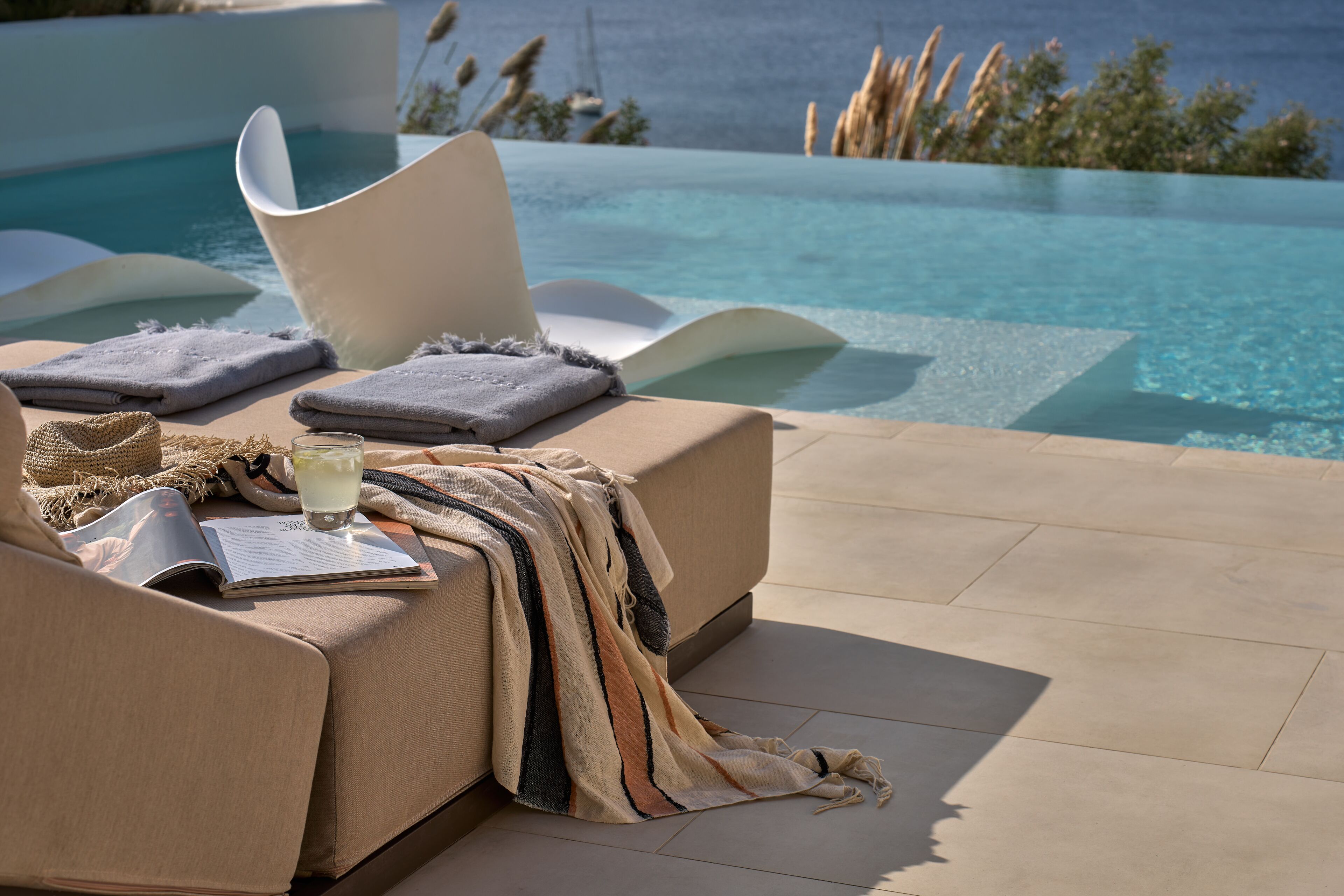 A sun lounger with a book and a glass of drink, overlooking a pool with clear water and a view of the sea.