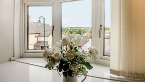 Ein Zimmer mit einem großen Fenster, das Buntglasdesigns zeigt, einer weißen Vase mit weißen Blumen und Blick auf Gebäude und Grünflächen.