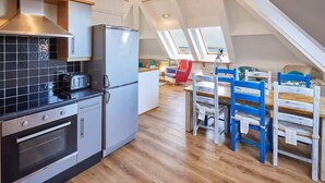 A modern kitchen with a built-in oven and a refrigerator, a dining area with a wooden table and blue chairs, and a loft bedroom with a skylight. - Host & Stay | The Watchtower (Waren Mill)