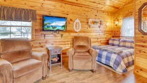 Ein gemütliches Blockhäuschen im Inneren mit Holzwänden, einem Fernseher, einem gepolsterten Sessel, einer Couch und einem Bett mit karierter Decke.