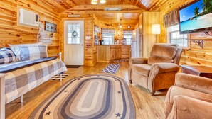Ein gemütliches Blockhaus-Interieur mit einem Bett, zwei Sesseln, einem Essbereich und einem Fernseher, der eine Strandansicht zeigt.