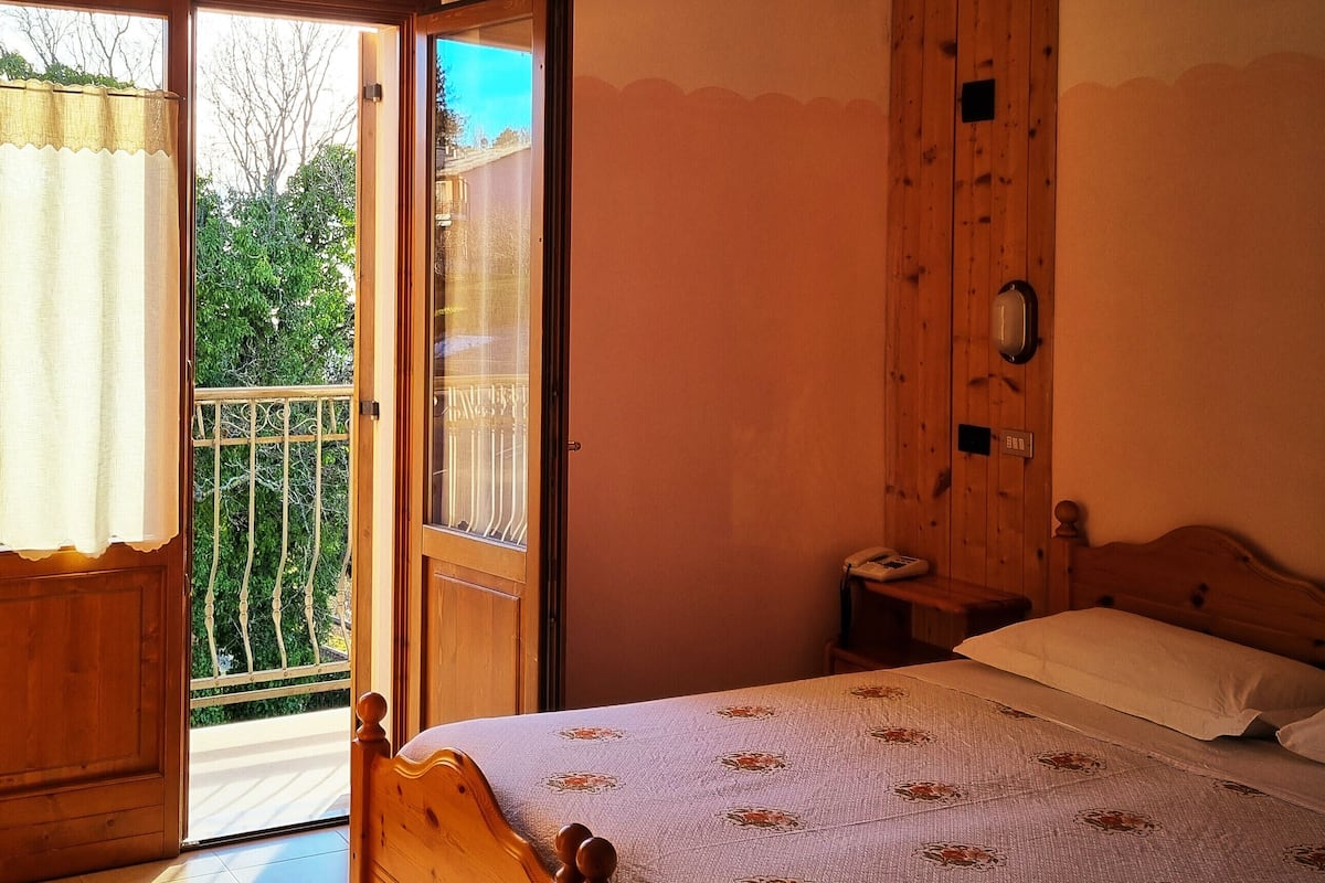 A bedroom with a wooden bed, a nightstand, a window with a view of greenery, and a door leading to an outdoor balcony.