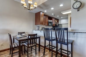 Uma sala de jantar com mesa e cadeiras de madeira, um candelabro e uma cozinha ao fundo.