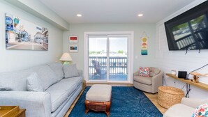 A living room with a grey sofa, a blue rug, a wicker ottoman, a television on the wall, and a balcony with a view. - 127th Bayside-pool-short walk 2 beach-By Park! (Ocean City)