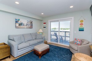 A living room with a grey sofa, a wooden side table, a blue rug, and a balcony with a view.