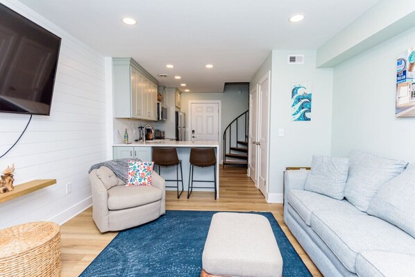 A living room with a sofa, ottoman, and a blue rug. There is a kitchen area with a dining table and chairs. - 127th Bayside-pool-short walk 2 beach-By Park! (Ocean City)