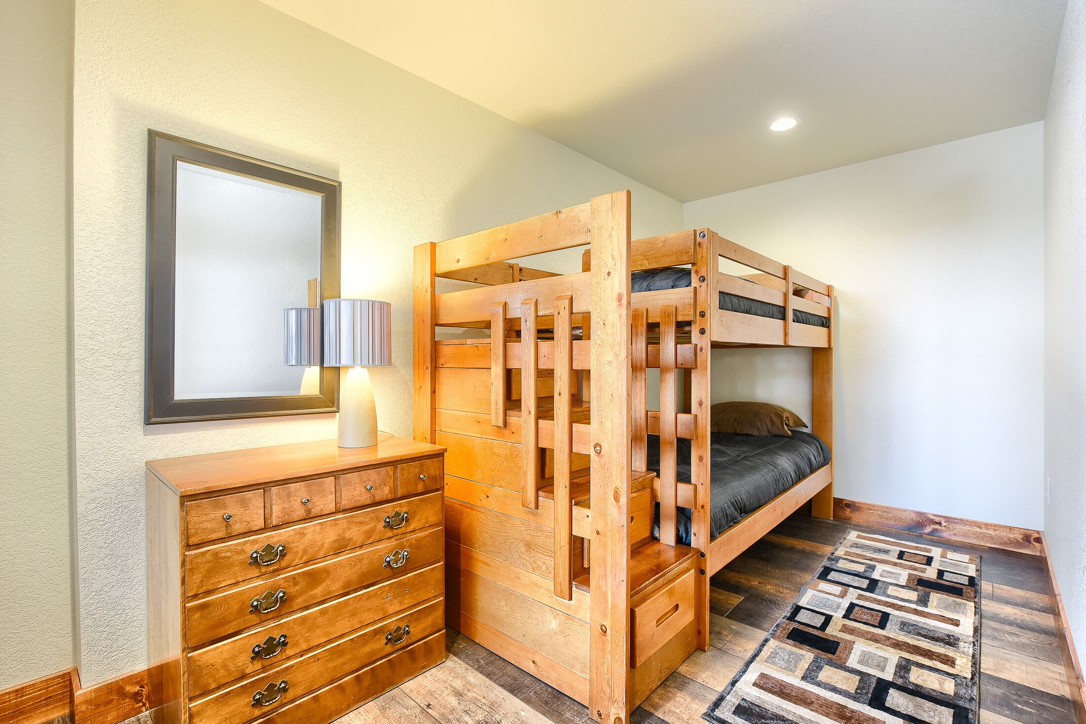 A bunk bed with a wooden frame, a dresser with drawers, a lamp, and a patterned rug on a wooden floor.