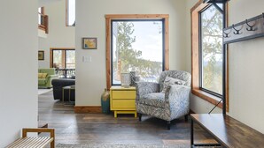 A cozy living room with a patterned armchair, a yellow side table, and a large window offering a view of trees. - New 3-Level Lodge w/ Amazing View Close to Slopes & Trails w/ Great Amenities (Lead)