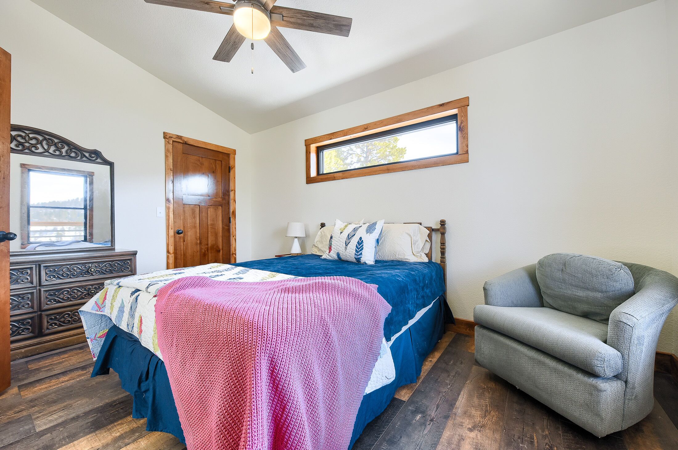 Un dormitorio con cama, una silla, cómoda, ventilador de techo y ventana.