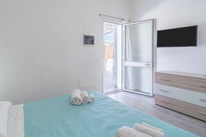 A hotel room with a bed, a TV mounted on the wall, a sliding glass door leading to an outdoor area, and a wooden dresser. - THE STACKS 3 (Lido di Noto)