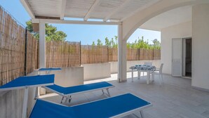 A covered outdoor area with two blue lounge chairs, a table with chairs, and a wooden fence in the background. - THE STACKS 3 (Lido di Noto)