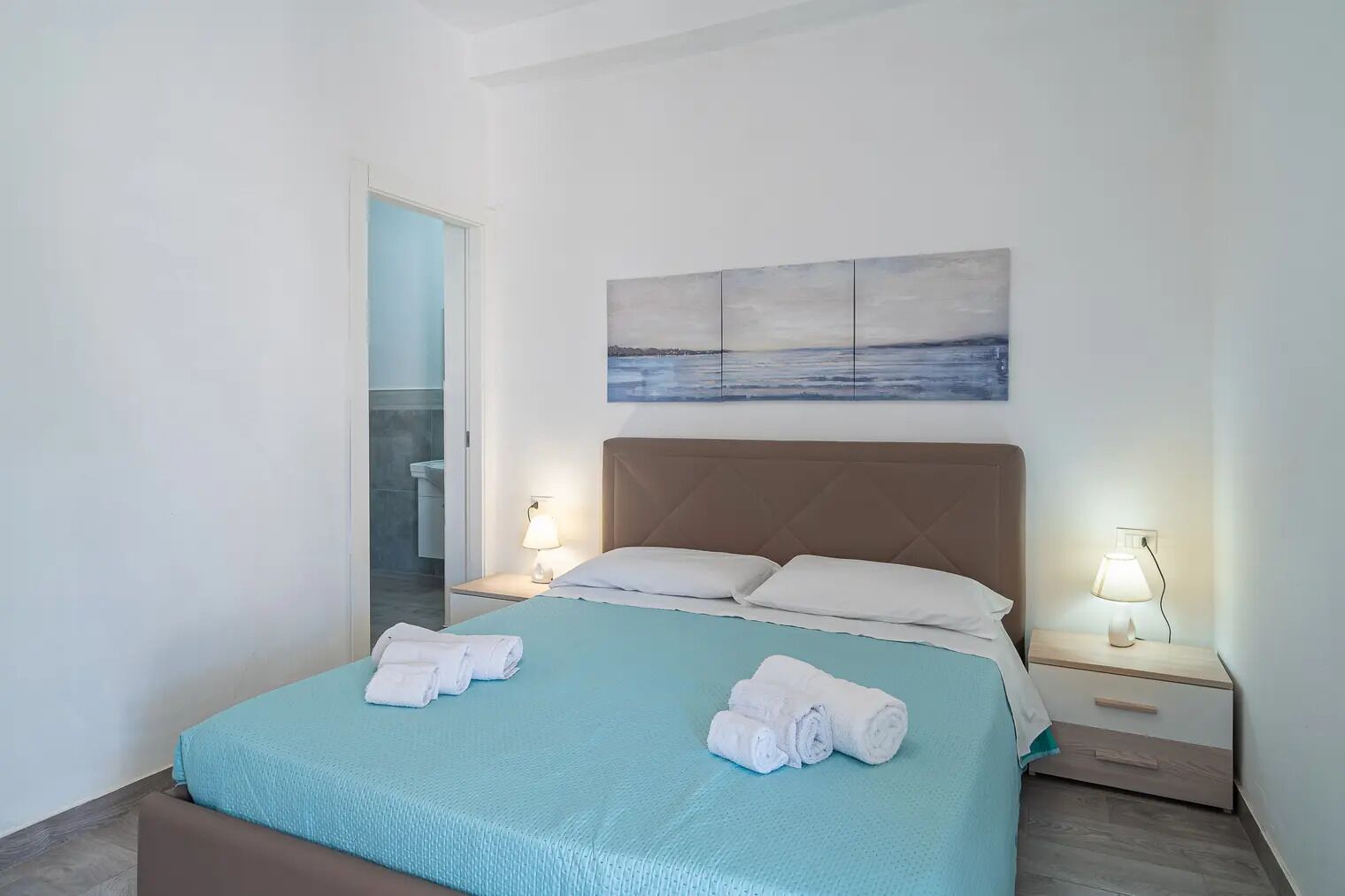 A neatly made bed with two white towels, a light blue bedspread, and a brown headboard. There is a painting of a seascape above the bed, and two bedside lamps are lit.