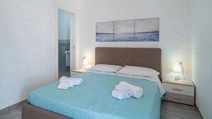 A neatly made bed with two white towels, a light blue bedspread, and a brown headboard. There is a painting of a seascape above the bed, and two bedside lamps are lit. - THE STACKS 3 (Lido di Noto)