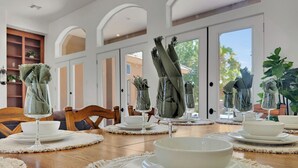 Un comedor con una mesa de madera dispuesta con platos, tazones y vasos blancos. Debajo de los utensilios hay protectores de mesa tejidos, y la mesa está ubicada frente a una puerta que da al exterior.