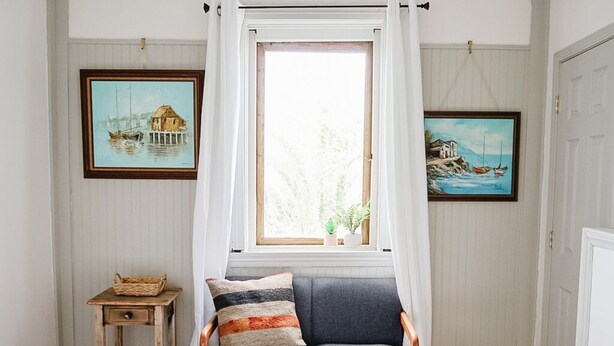 Une chambre à coucher avec un lit, une fenêtre ornée de rideaux blancs, un fauteuil avec des coussins et deux tableaux encadrés accrochés aux murs.