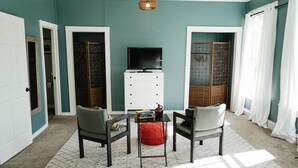 Un salon aux murs de couleur turquoise, avec un meuble blanc, deux fauteuils, un pouf rouge et un téléviseur posé sur un meuble.