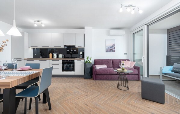 Eine moderne Küche mit weißen Schränken, ein Essbereich mit einem Holztisch und Stühlen, ein violettes Sofa und ein kleiner runder Tisch mit Flasche und Glas.