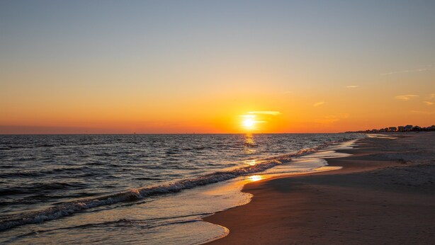 Una playa al atardecer, con el sol ocultándose en el horizonte, mar tranquilo y una orilla arenosa.