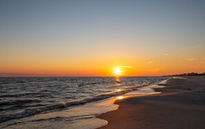 Una playa al atardecer, con el sol ocultándose en el horizonte, mar tranquilo y una orilla arenosa.