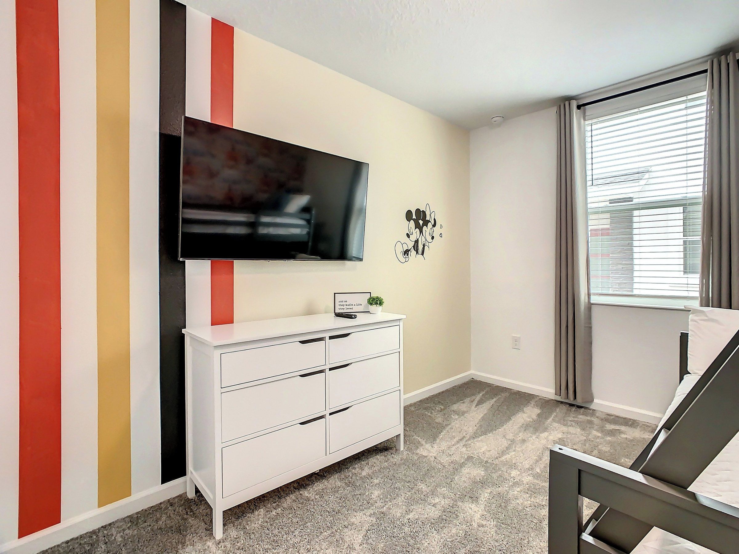 Quarto com cama, televisão fixada na parede, penteadeira e janela com cortinas.