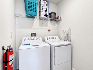 A laundry room with two front-loading washing machines, a fire extinguisher, and a shelf with cleaning supplies.