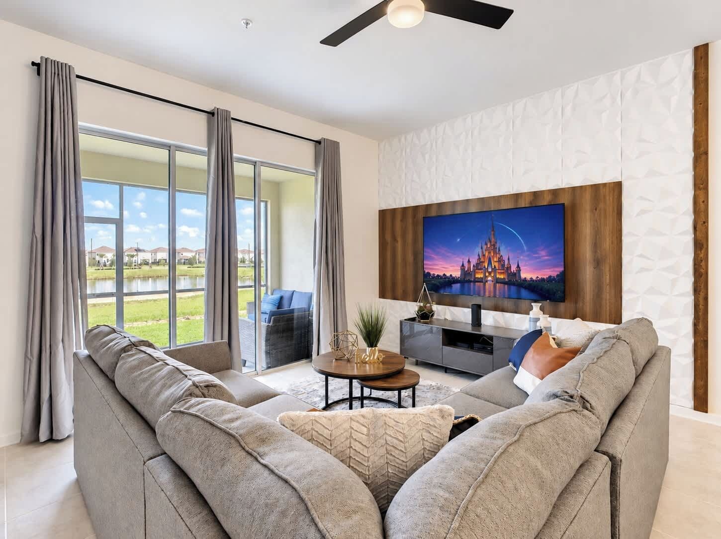 Uma sala de estar moderna com uma grande TV de tela plana fixada em uma parede texturizada, um sofá cinza e vista para um lago através de janelas amplas.