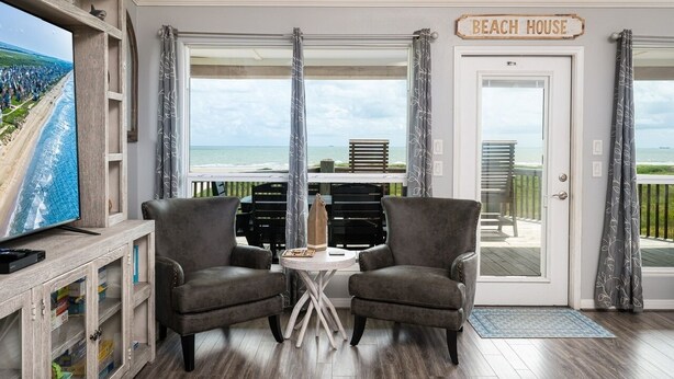 Ett rum med en TV, två fåtöljer, ett litet bord, en dörr med texten ”STRANDHUS” och utsikt över stranden.