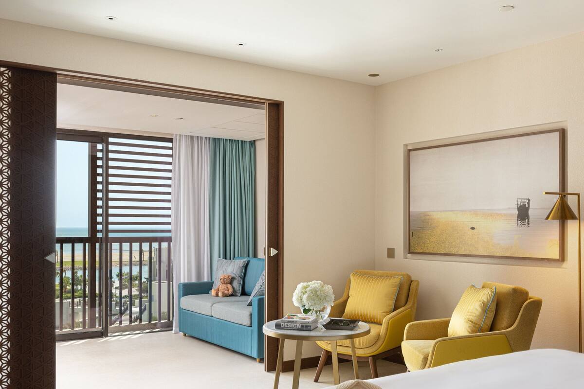 A hotel room with a large bed, a sofa, a chair, a small table, and a balcony with curtains.