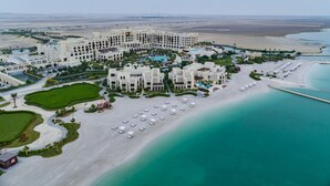 An aerial view of a resort with multiple buildings, a beach, and a pool area. - Jumeirah Gulf of Bahrain (Zallaq)